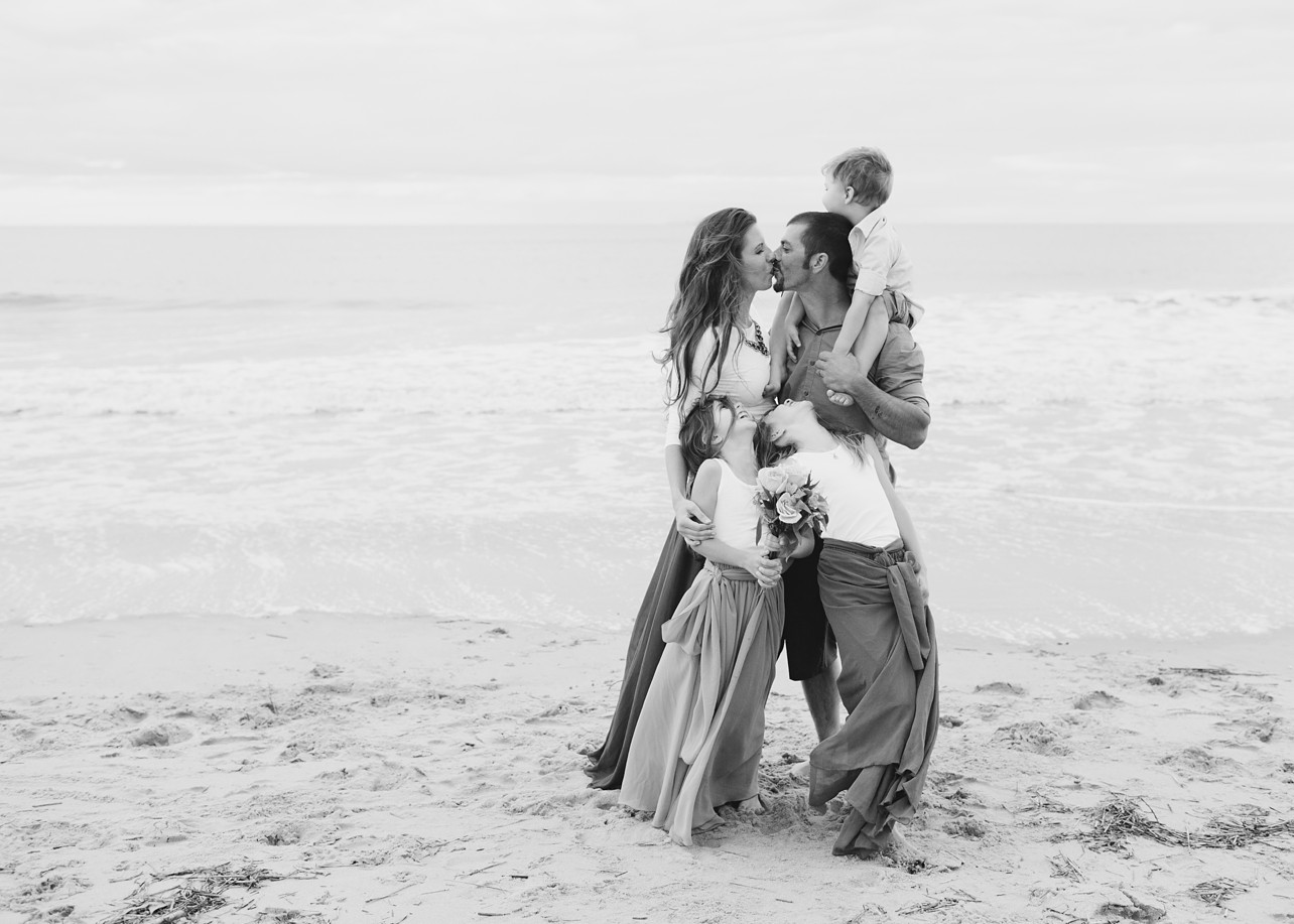 mother and father with son on his shoulder, kiss on the beach while their daughters smile up at them.