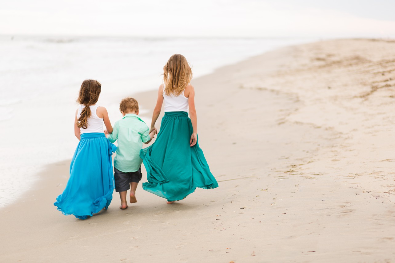 Three children walk along the beach holding hands, their backs to the camera. 