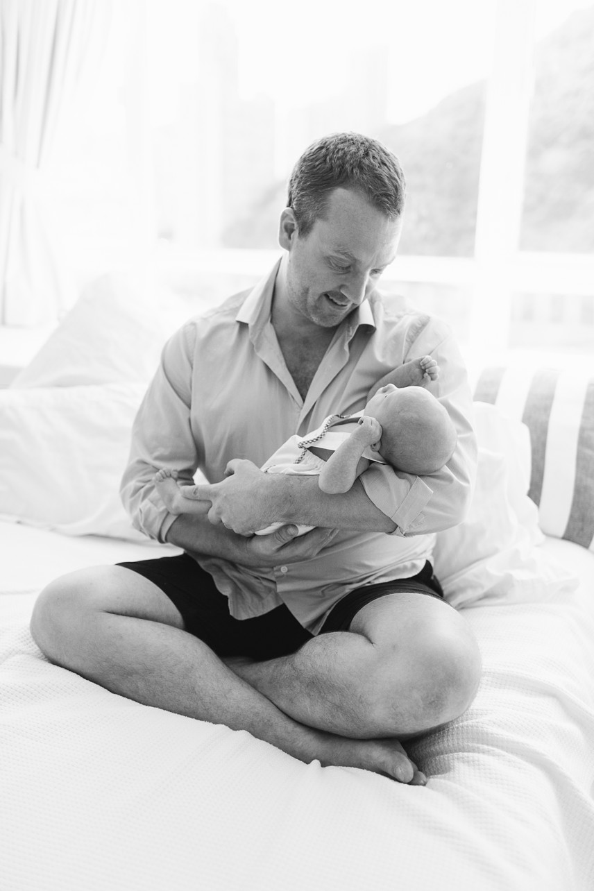 black and white photo of father sitting on their bed holding his newborn baby son. 