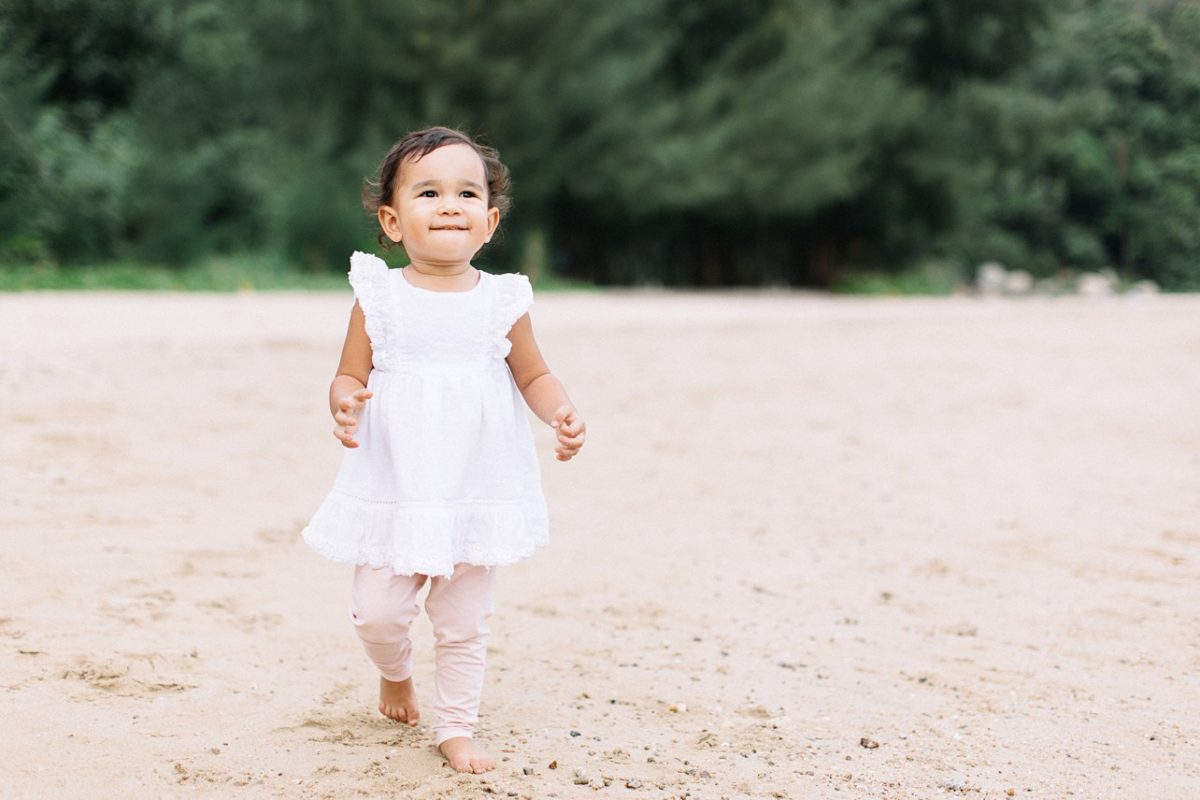 a toddler in white dress and pink pants smiling on the beach