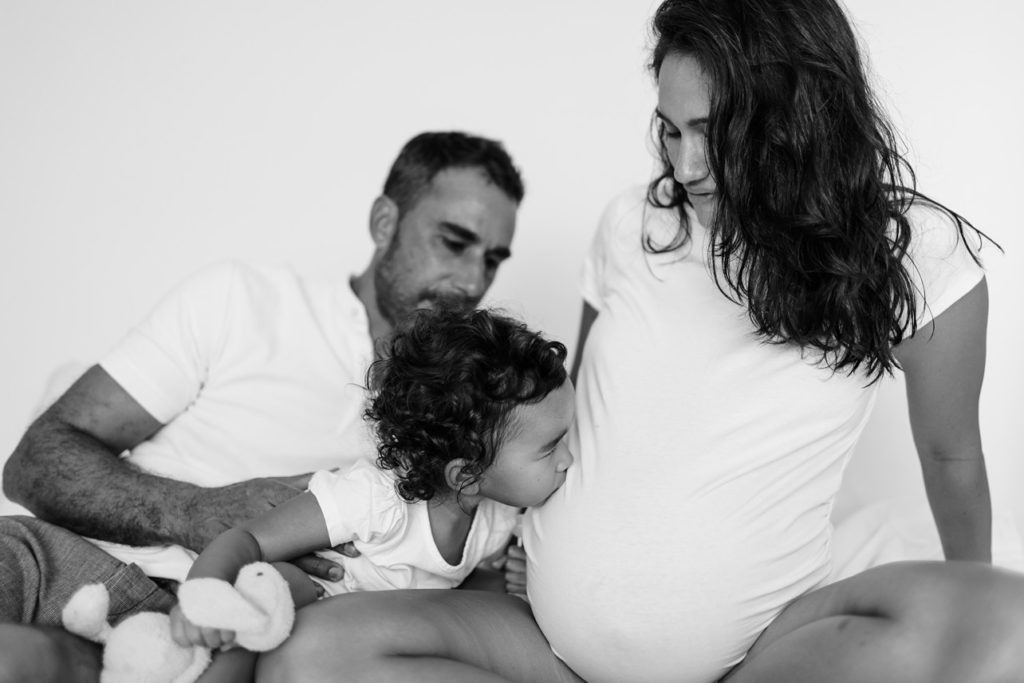 family of 3, mom is pregnant and little girl who sits in front of her dad is kissing the belly in black and white image