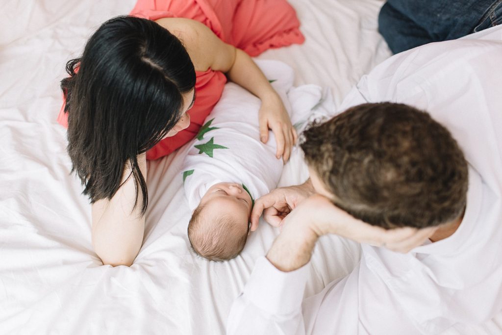 new family of 3 with newborn baby are laying down on the bed, mom is in pink dress