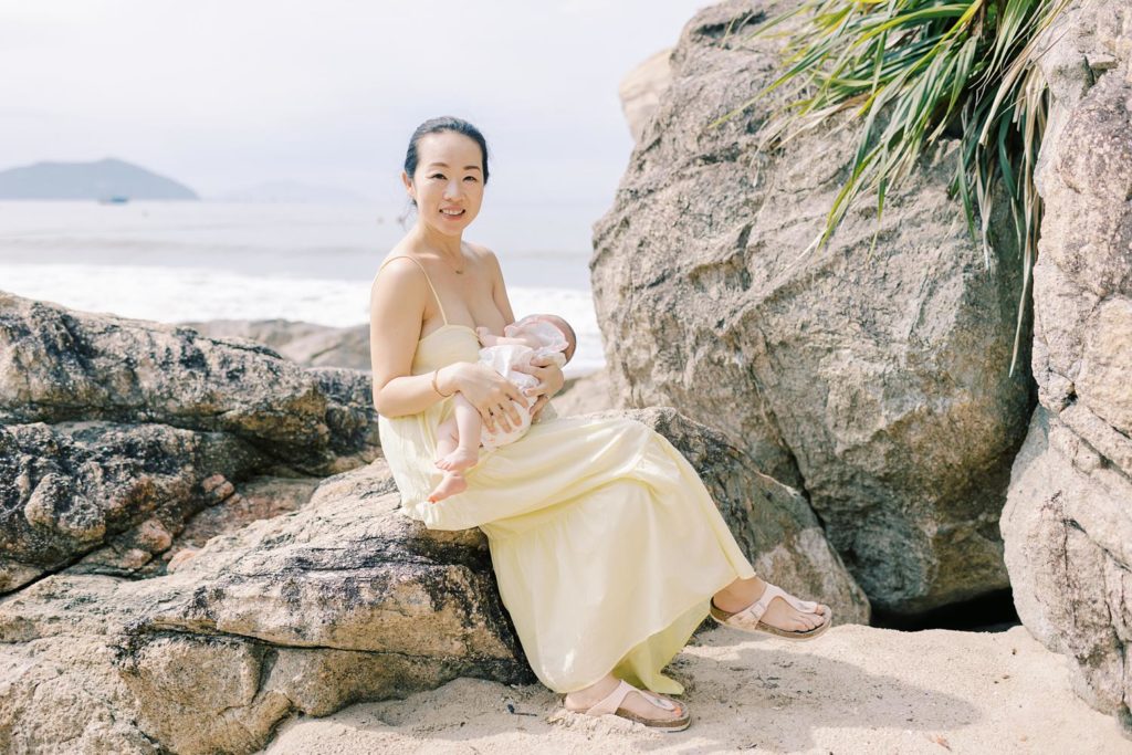 Smiling mother in a yellow dress, sitting on a rock by the beach, breastfeeding her baby.