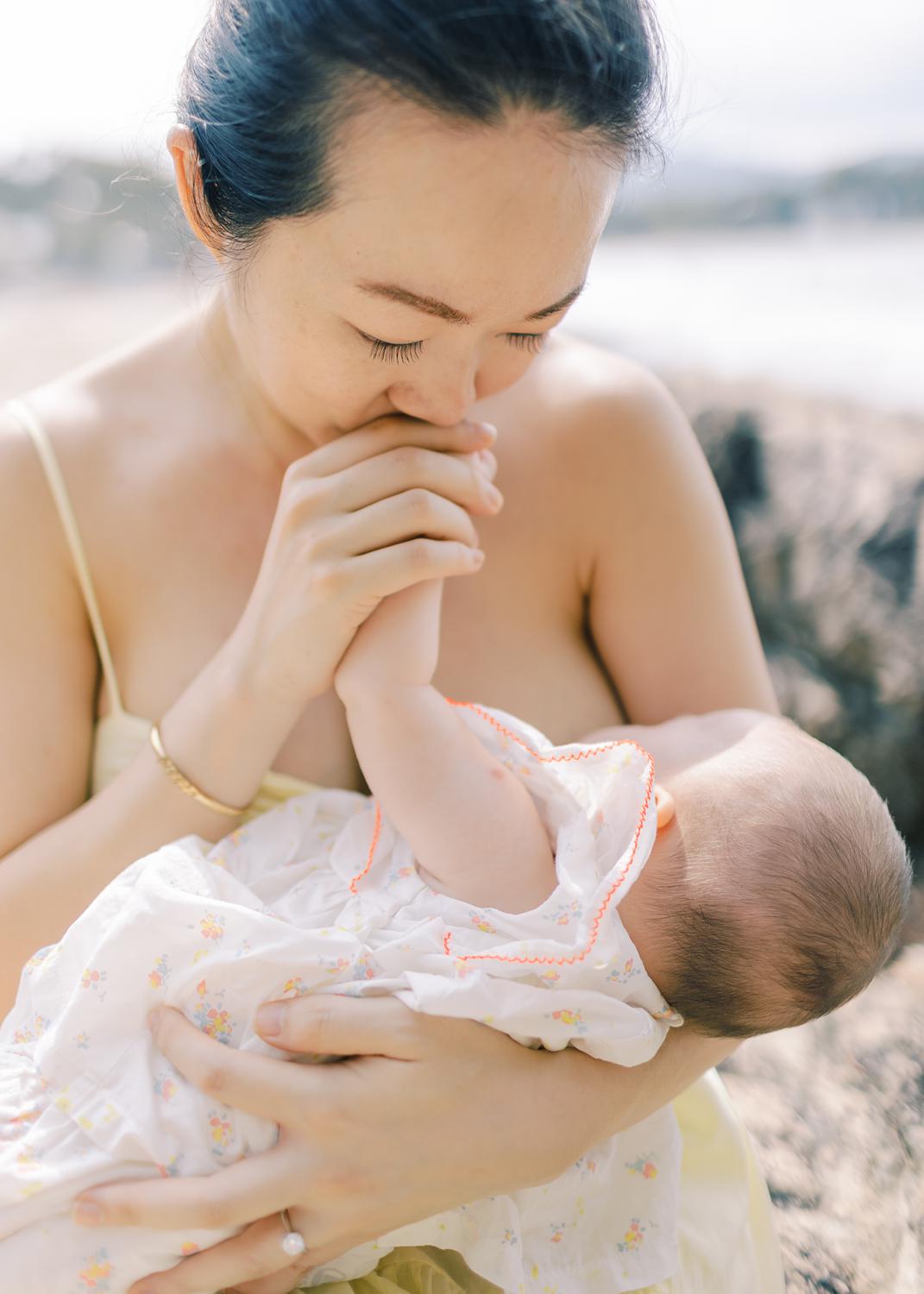 Mother is kissing baby's hand while she breastfeeds her baby girl during their nursing photoshoot.

