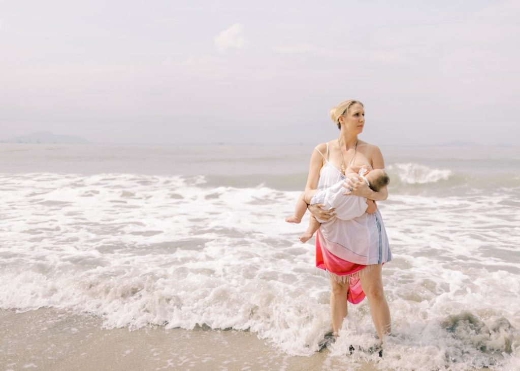 a mother in beautiful white and pink dress is proudly breastfeeding her daughter on the shore at the beach