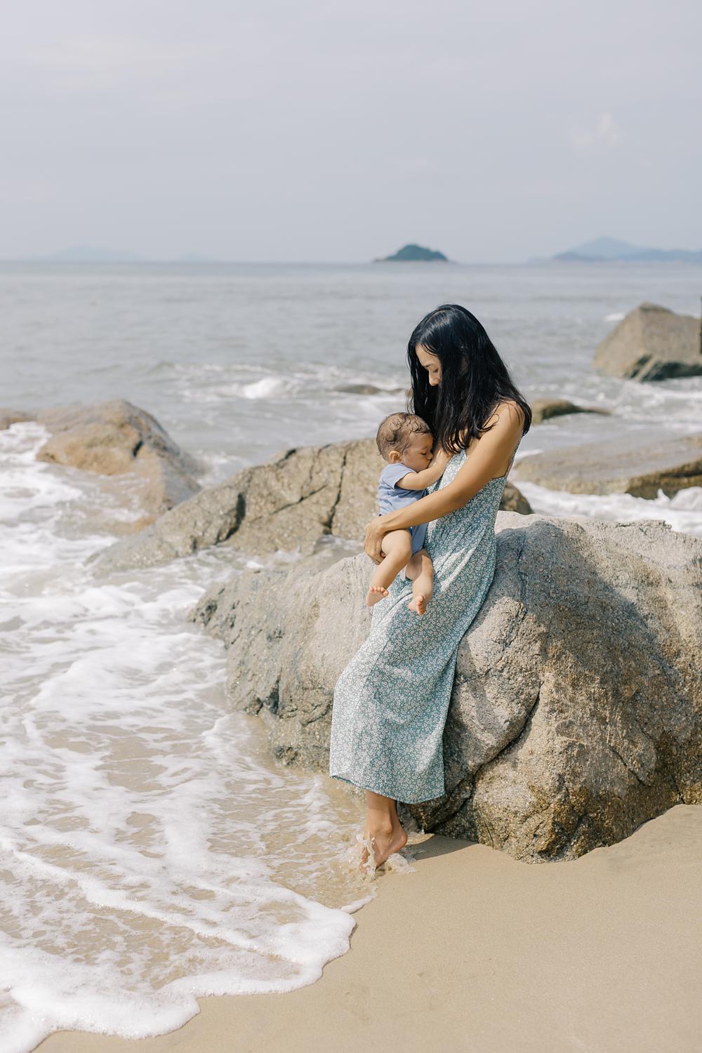 mother in blue floral dress is breatfeeding her son leaning on the huge rock on the beach